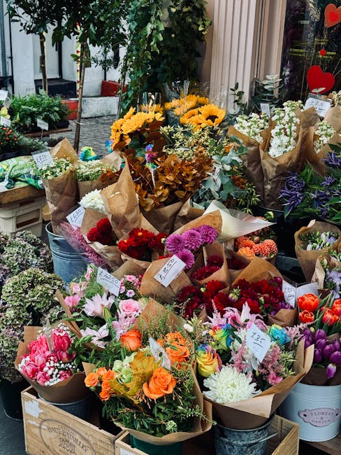 A vibrant display of freshly arranged flower bouquets outdoors on a shop display stand. The arrangements include a variety of flowers such as bright yellow sunflowers, pink and orange roses, purple ir