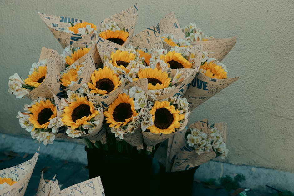 A vibrant bouquet of fresh sunflowers with large yellow petals and dark brown centers, wrapped in brown paper printed with blue and black words such as 'love' and 'happy.' The bouquet is arranged in a