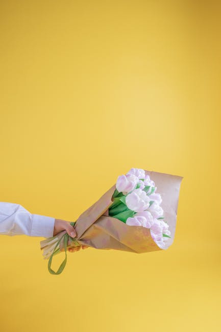 A hand holding a bouquet of fresh light pink tulips wrapped in brown craft paper with green stems visible, presenting a soft and delicate floral gift against a vibrant yellow background. The tulips ar