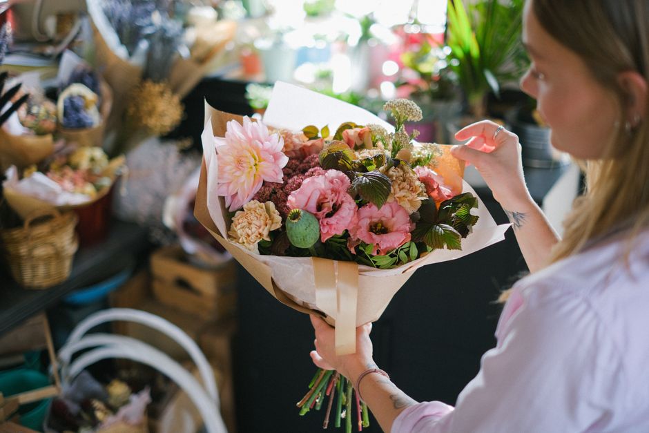 A woman holding a fresh floral bouquet wrapped in light brown paper and tied with a beige ribbon, featuring pink dahlias, light pink roses, deep green foliage, and small white accent flowers. She is s